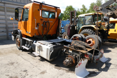 Orange Volvo truck cab with chassis and attached drivetrain components in an outdoor lot, other heavy equipment visible in background.