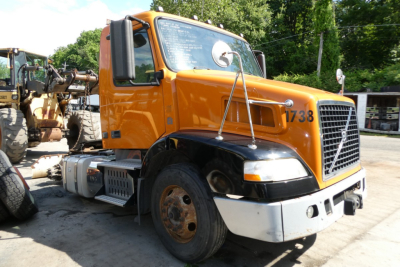 Orange Volvo VNM42T conventional daycab truck, front-right view, with large grille, black fenders, white bumper, and worn front left tire; VIN tag visible.