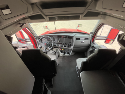 Kenworth T680 cab interior with dashboard and steering wheel, view from rear seating area; red exterior visible through windshield.