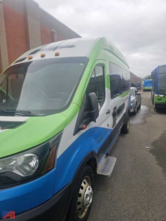 2019 Ford Transit full-size van in a multi-color livery (green, blue, white) parked in a lot; side view shows front door, wheel, and running boards.