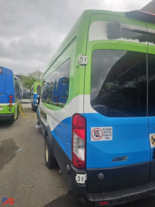 Ford Transit full-size van rear side view showing green/blue/white livery, rear door, taillight, and bumper.