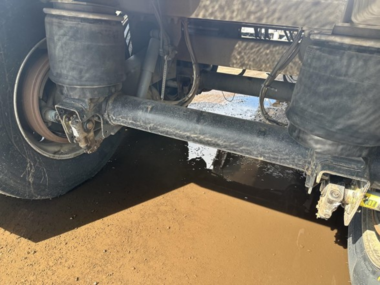 Trailer underside showing solid steel axle and suspension components with a wheel on left, hydraulic lines, and a muddy ground surface.