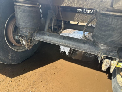 Trailer underside showing solid steel axle and suspension components with a wheel on left, hydraulic lines, and a muddy ground surface.