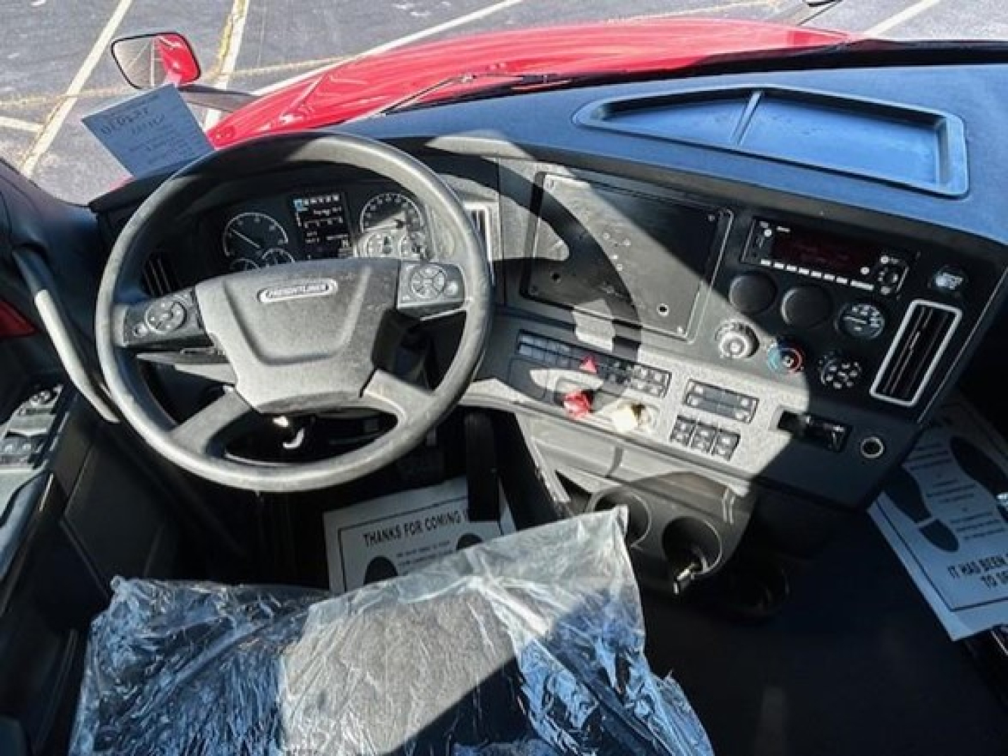 Cab interior of a 2023 Freightliner Cascadia 126 showing steering wheel, dashboard gauges, center console, and plastic-covered seat.
