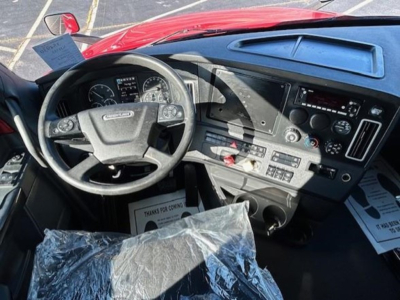 Cab interior of a 2023 Freightliner Cascadia 126 showing steering wheel, dashboard gauges, center console, and plastic-covered seat.