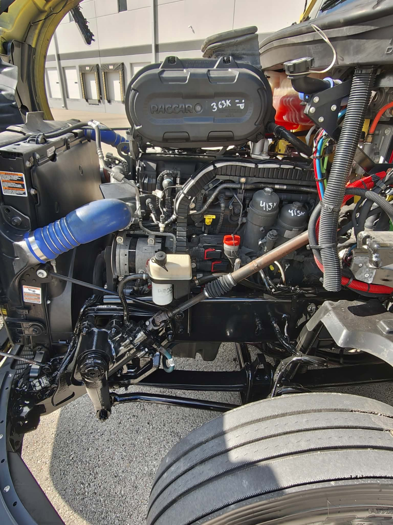 Kenworth T680 engine bay with exposed components including air intake, hoses, wiring, and turbocharger visible; view from left front shows engine and front wheel.