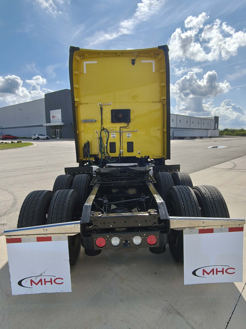 Rear view of yellow Kenworth T680 sleeper truck cab with dual rear wheels, mudflaps labeled MHC, and rear chassis wiring. Visible text includes 'MHC' on mudflaps and yellow cab back.