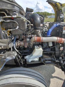 Kenworth T680 engine bay showing coolant reservoir, intercooler pipes, hoses, and turbocharger components in a daytime service area.