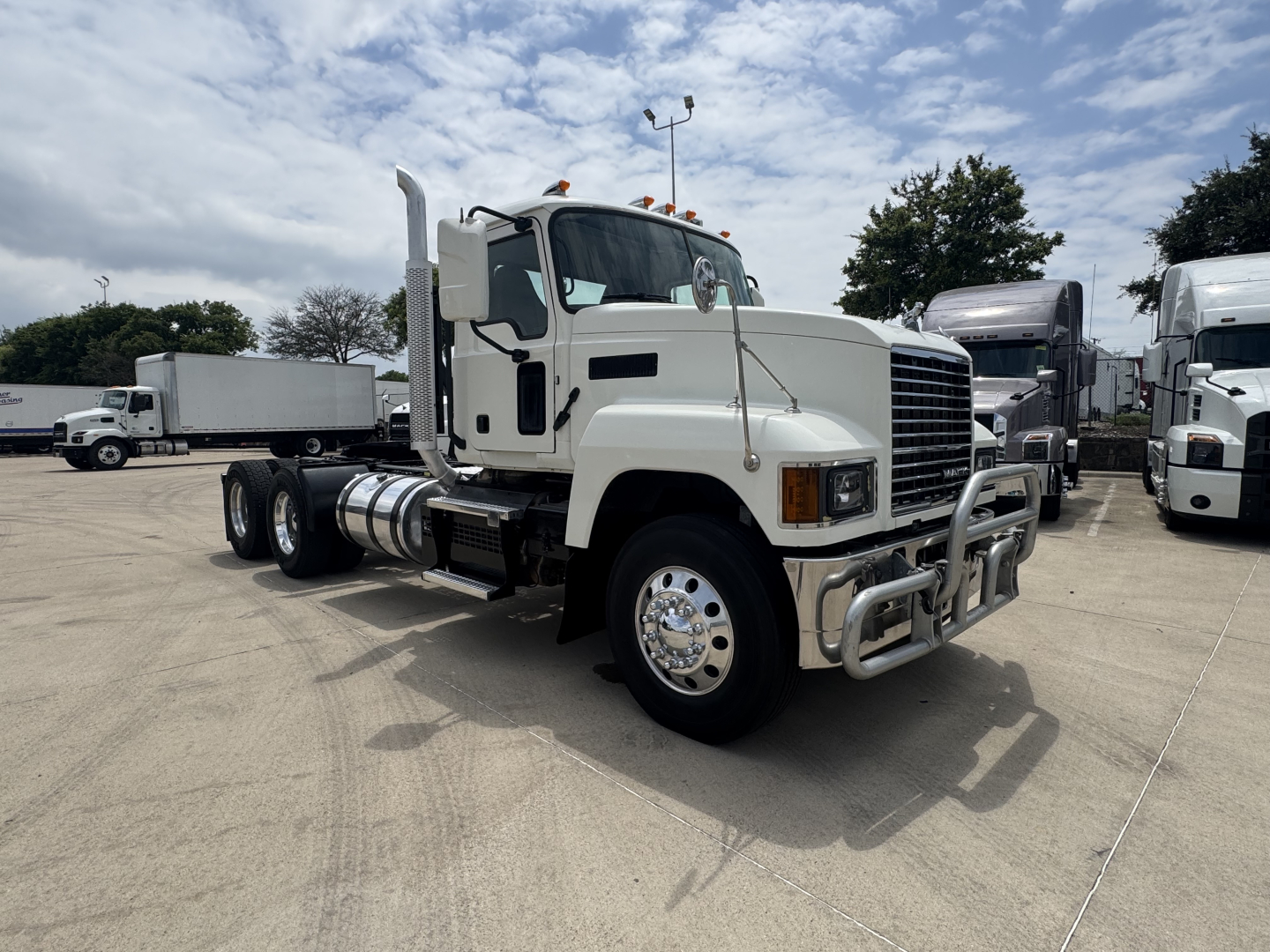 Truck cab and chassis showing front grille with bumper; Mack PI64T in a light color with dual rear axles and side fuel tanks. Visible text includes no specific model labels on the cab; equipment appears in a dealership lot.
