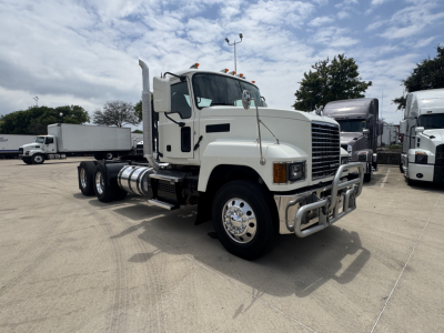 Truck cab and chassis showing front grille with bumper; Mack PI64T in a light color with dual rear axles and side fuel tanks. Visible text includes no specific model labels on the cab; equipment appears in a dealership lot.