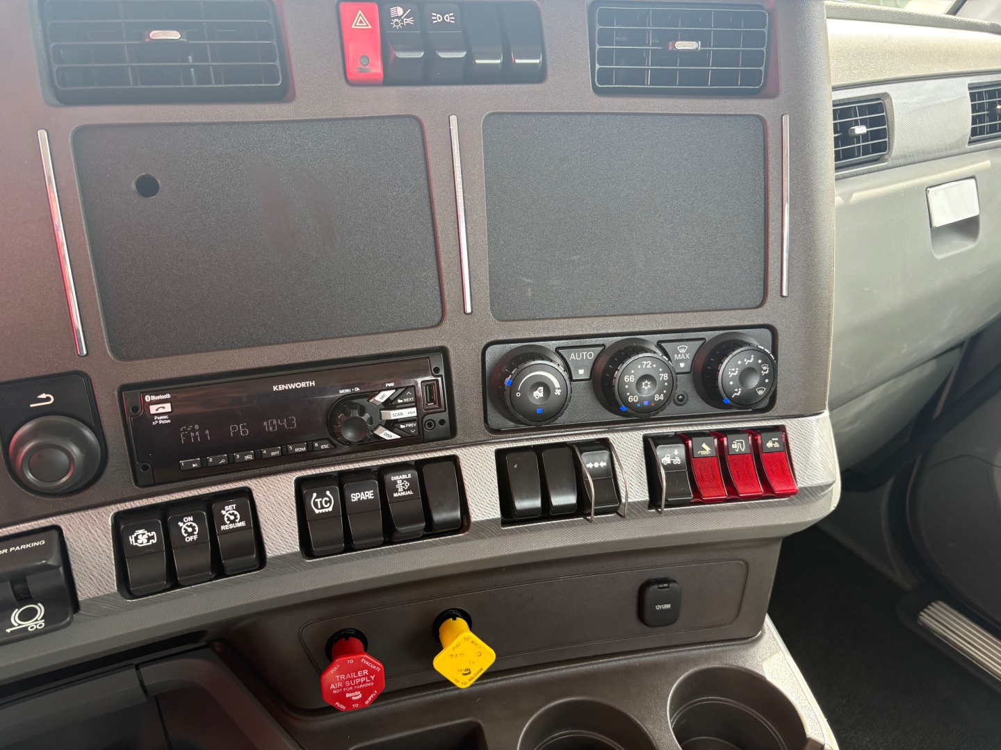 Panel view of 2019 Kenworth T880 dashboard showing climate controls, radio, multiple switches, and two colored knobs; red and yellow icons for controls visible.
