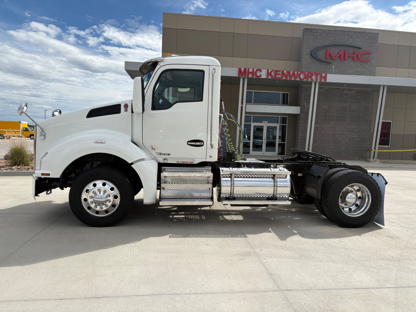 Kenworth T880 conventional daycab truck side view showing chassis, fuel tanks, and cab; dealership building in background