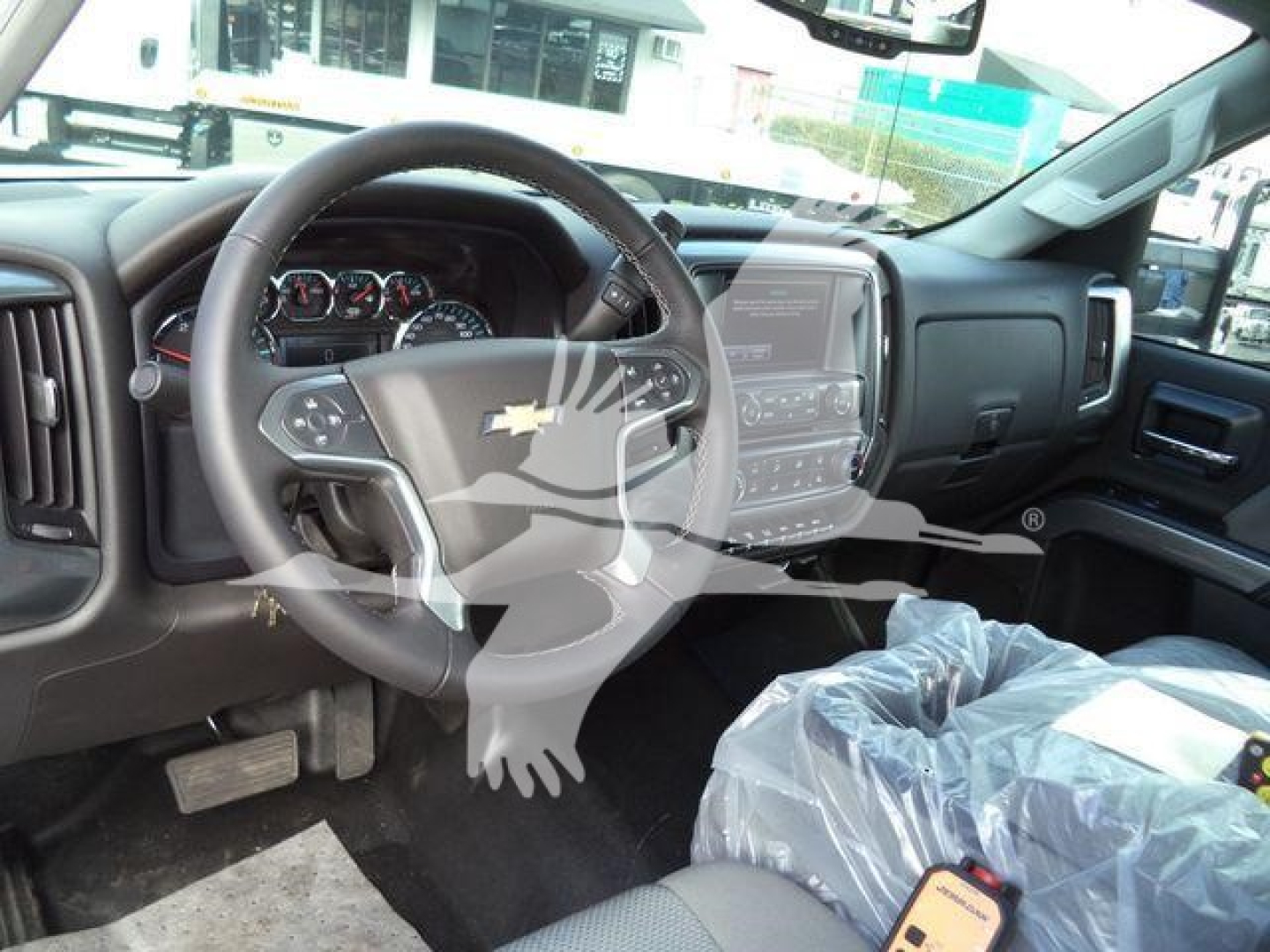 Truck interior showing steering wheel with Chevrolet logo, dashboard gauges, central touchscreen, and plastic-covered passenger seat; visible stock label reflections in windshield.