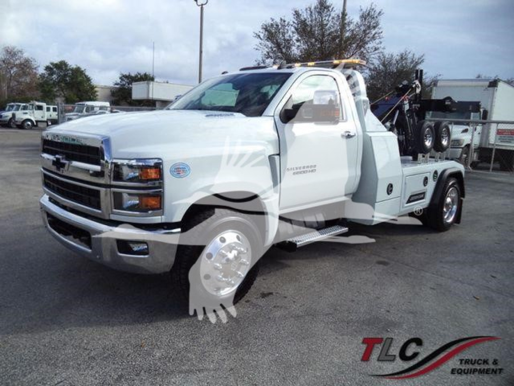 Truck image showing a white 2023 Chevrolet Silverado 6500HD tow truck with a wheel-lift bed, winch system, and towing equipment.
