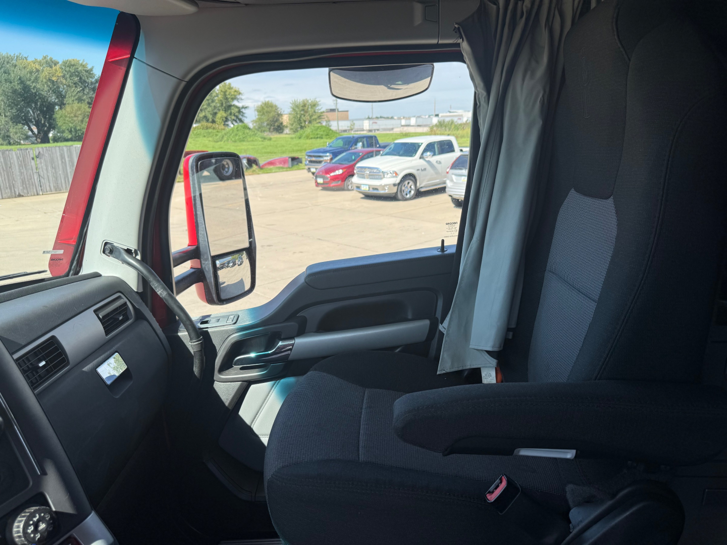 Truck cab interior view with steering wheel, side window, and seat; outside shows other vehicles in a lot.