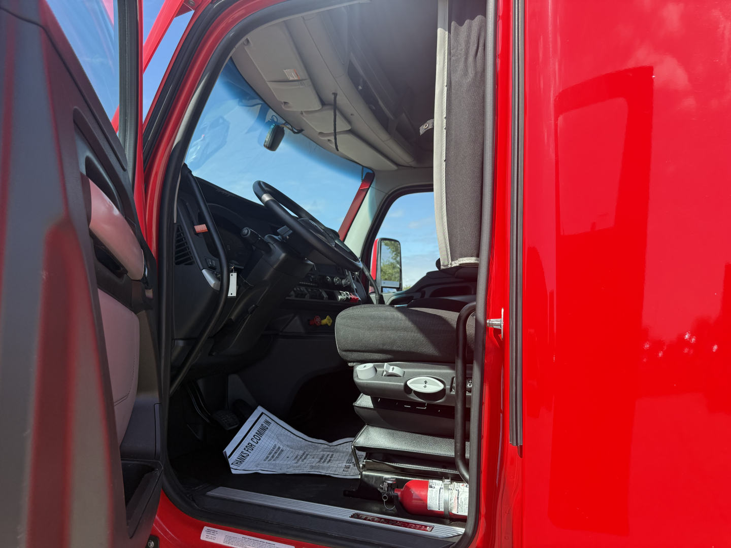 Kenworth T680 truck cab interior view from open driver door showing steering wheel, dashboard, driver's seat, and red exterior panel.