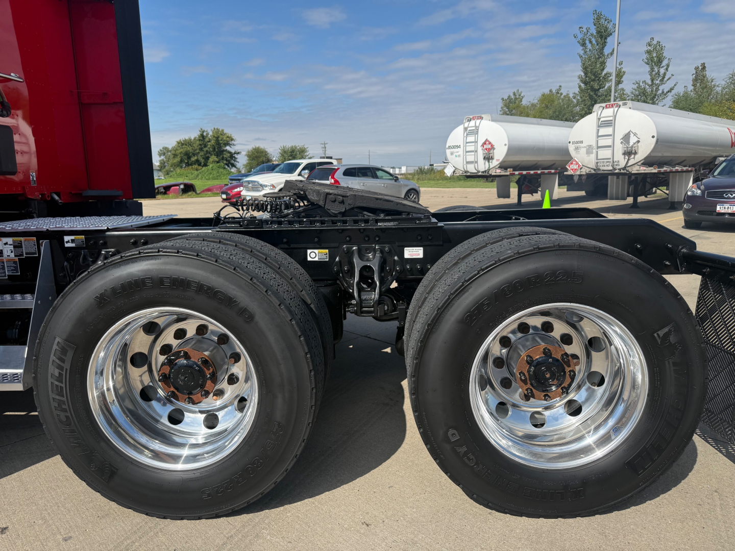 Dual rear axle on a Kenworth truck chassis with chrome wheels and black frame, fuel tanks and trailers in background.