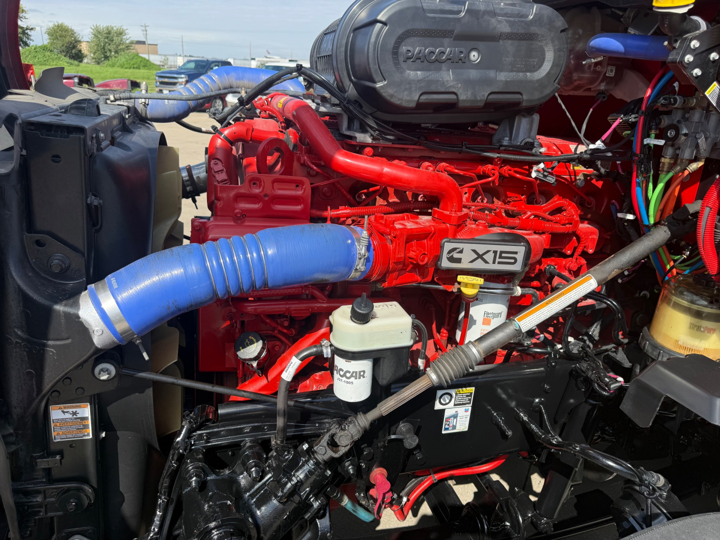 Kenworth T680 engine bay showing red engine with transverse turbo piping, blue intercooler hose, coolant reservoir, and PACCAR components.