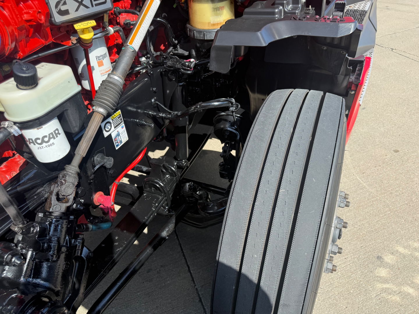 Kenworth T680 engine bay with visible fluid reservoirs and front suspension; shows coolant reservoir, power steering/other fluid lines, and part of the stacked front tires.