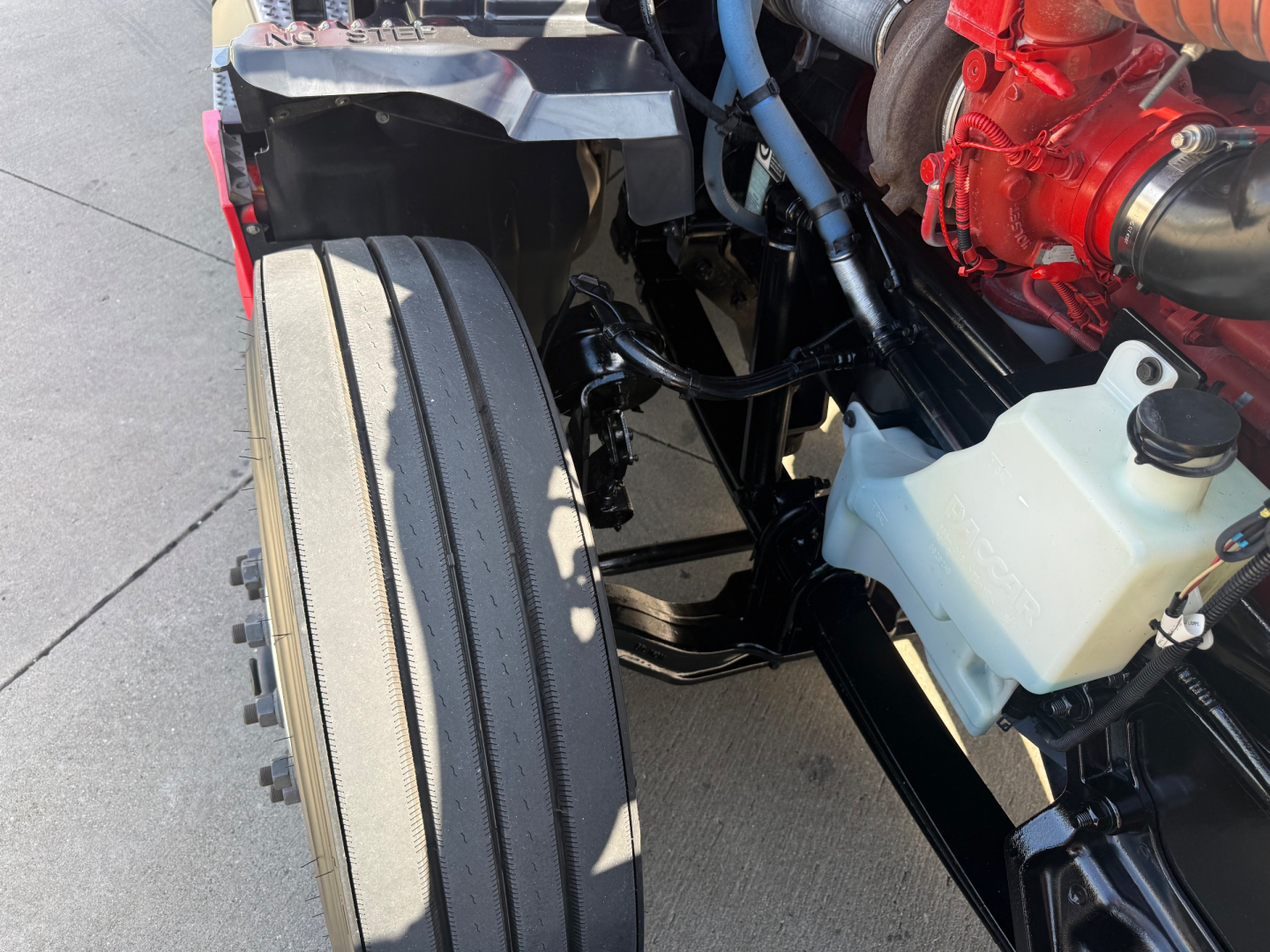 Front left wheel and engine bay of a 2023 Kenworth T680 showing part of the coolant reservoir, red engine component, and black chassis.