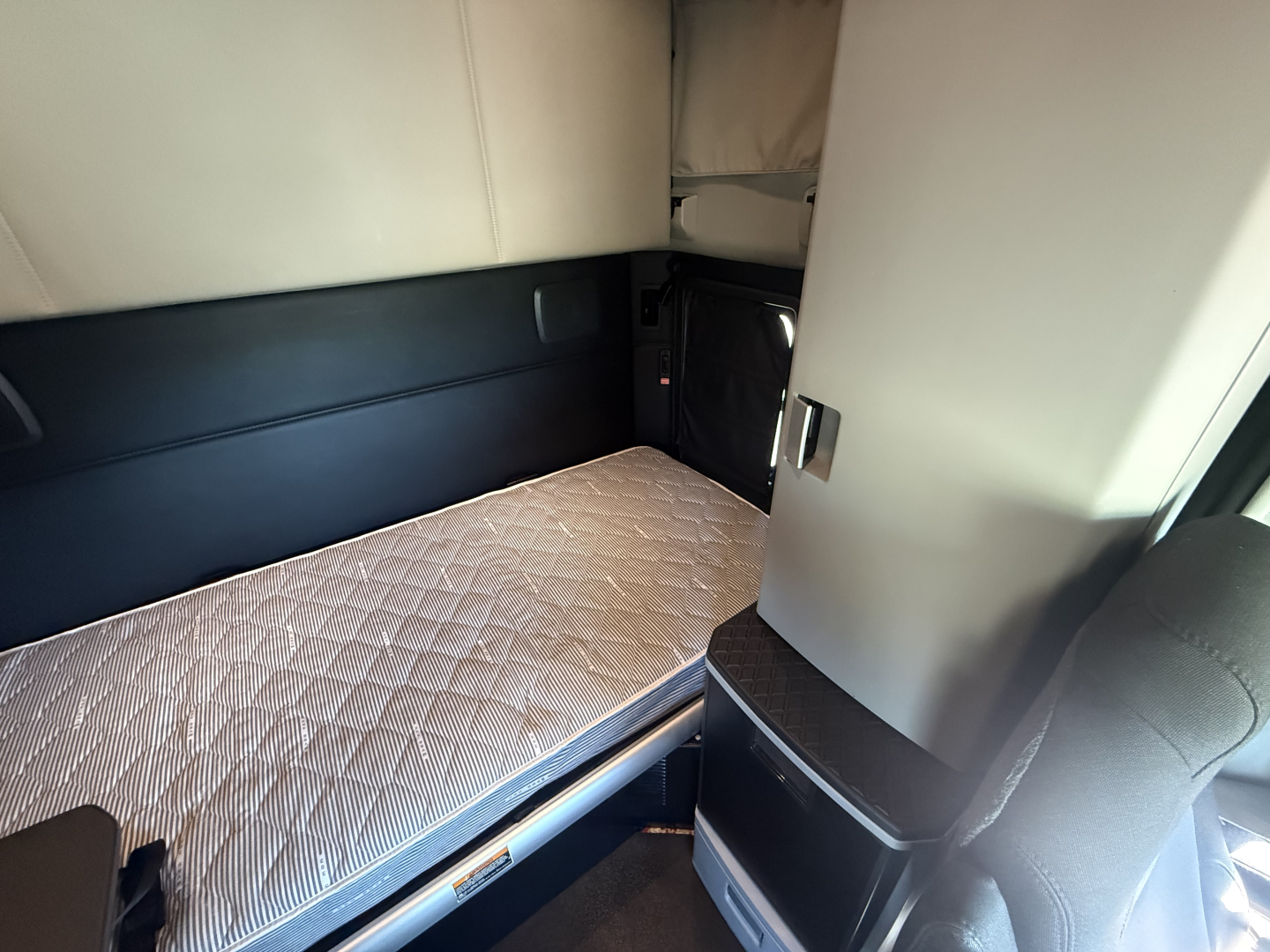 Kenworth T680 sleeper cab interior with a single bunk and quilted mattress, beige and black upholstery, storage compartments, and a small window in the sleeper area.
