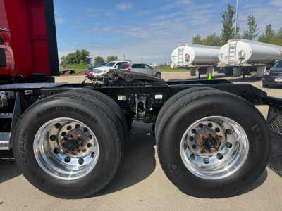 Dual rear axle on a Kenworth truck chassis with chrome wheels and black frame, fuel tanks and trailers in background.
