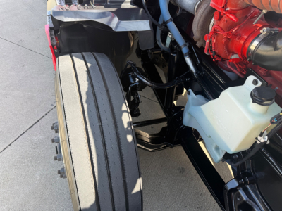 Front left wheel and engine bay of a 2023 Kenworth T680 showing part of the coolant reservoir, red engine component, and black chassis.
