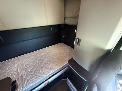 Kenworth T680 sleeper cab interior with a single bunk and quilted mattress, beige and black upholstery, storage compartments, and a small window in the sleeper area.