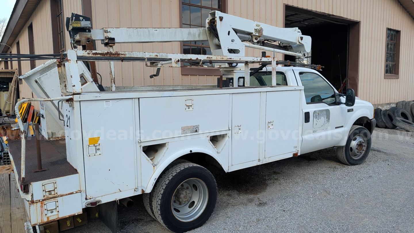 Ford F-550 bucket truck side view showing service utility bed with compartments and hydraulic boom on top, white utility truck, gravel yard, storage building in background.