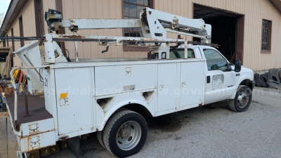 Ford F-550 bucket truck side view showing service utility bed with compartments and hydraulic boom on top, white utility truck, gravel yard, storage building in background.