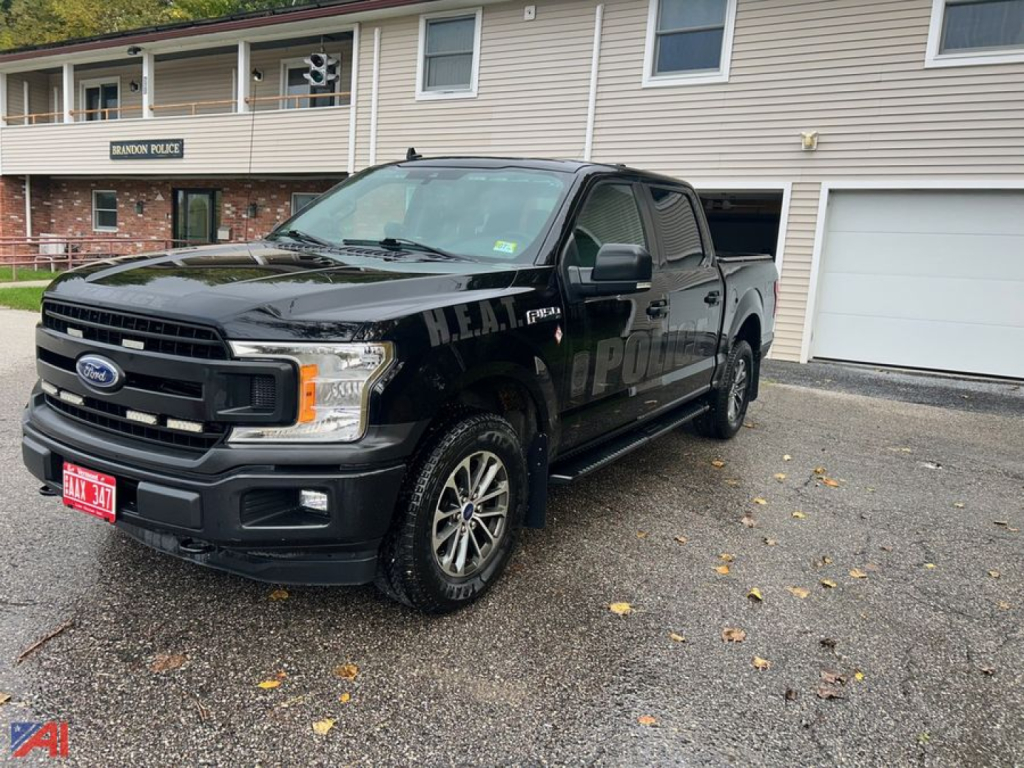 Ford F-150 police package vehicle 2020 crew cab 4x4 in black with HEA? markings on doors, front grille, red license plate, parked in lot