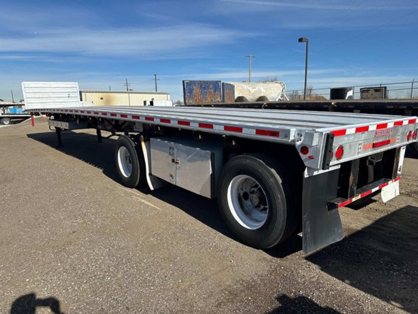 Flatbed trailer from the side showing dual rear wheels and storage box with metal toolbox and reflective red-and-white safety tape along the rails.