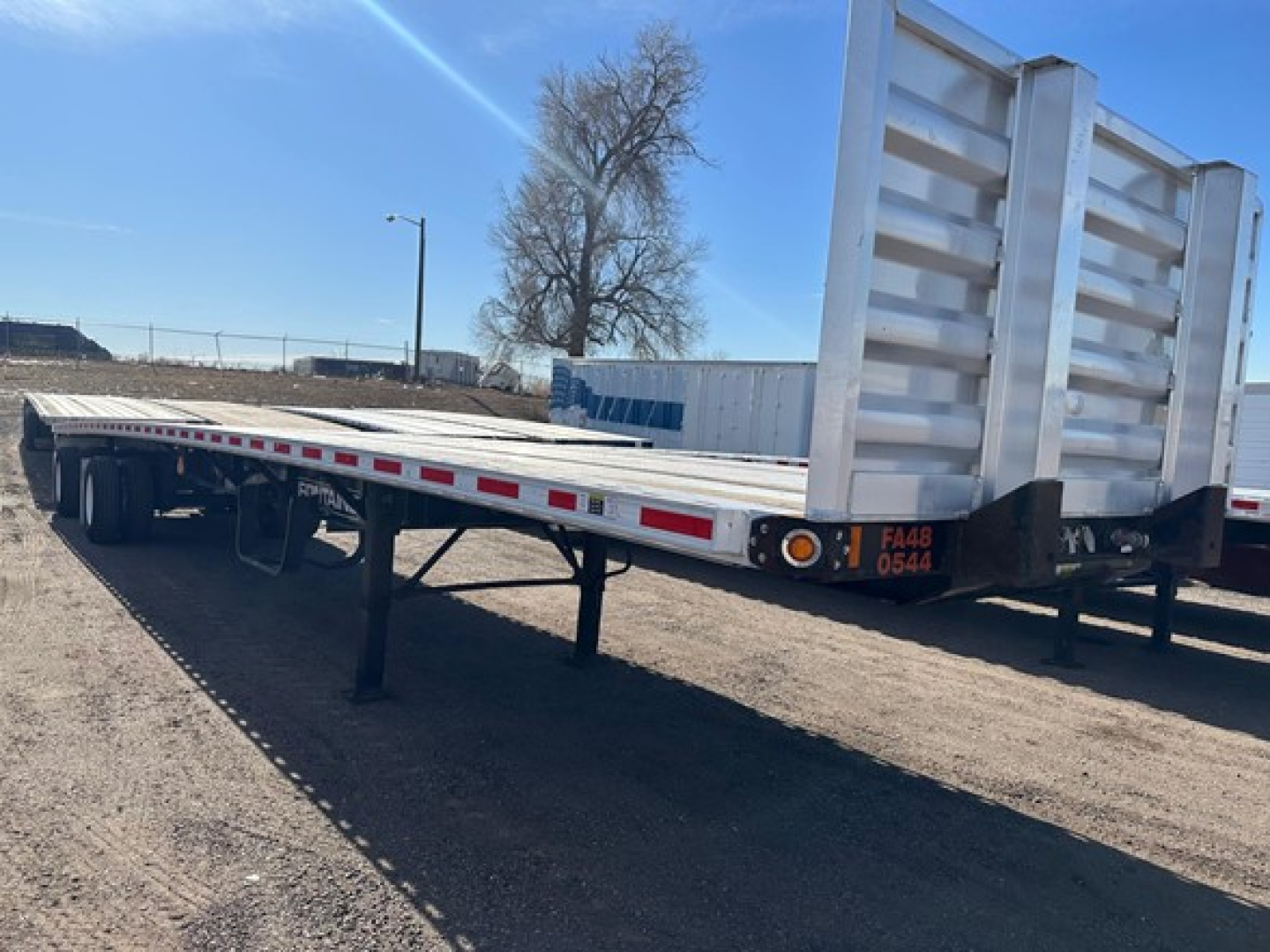 Flatbed trailer with low angle view, Fontaine Infinity branding not visible, two-wheel dual axles, long steel flatbed with rear riser gate, FCC/serial-looking tag near corner.