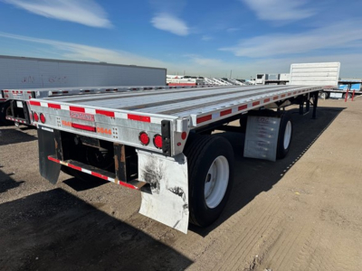 Flatbed trailer in a yard showing tandem axles and reflective red/white safety stripes along the sides, rear with mud flaps and taillights.
