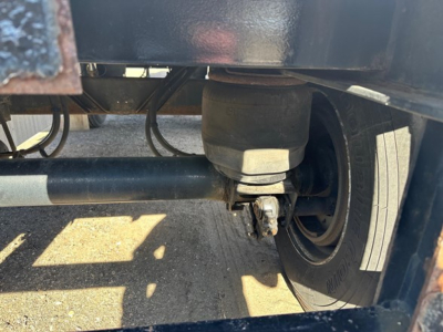 Trailer undercarriage showing axle with leaf spring, wheel, and tire. Visible components include a cylindrical axle tube, suspension mount, and brake linkage.