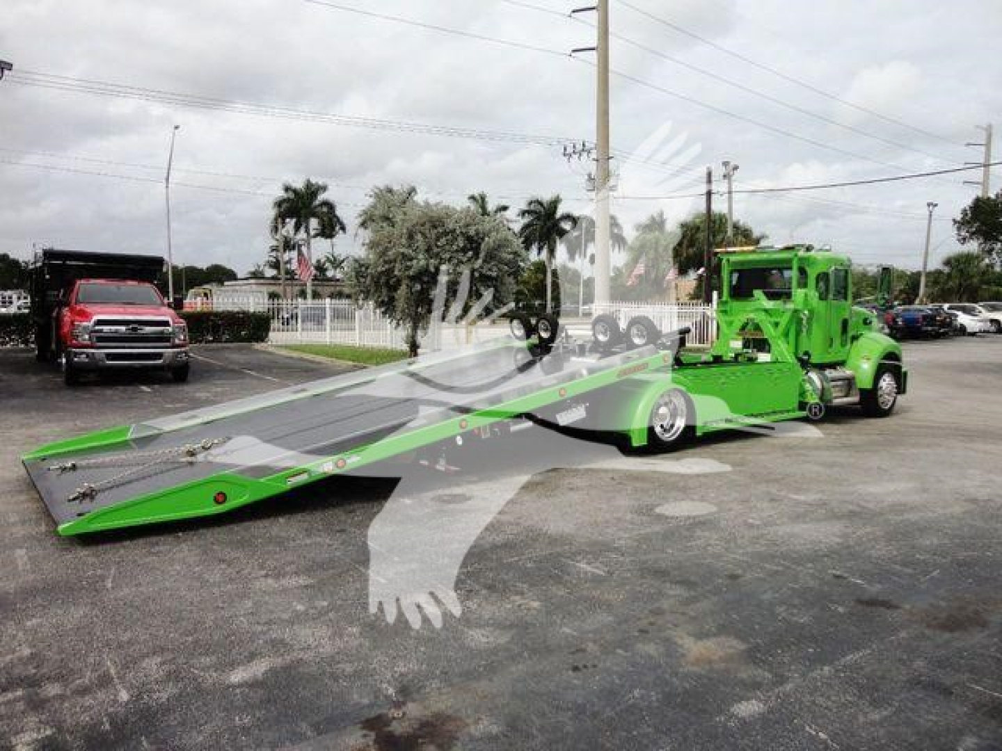 Green Peterbilt rollback tow truck with extended flatbed and cab; other vehicles in background.