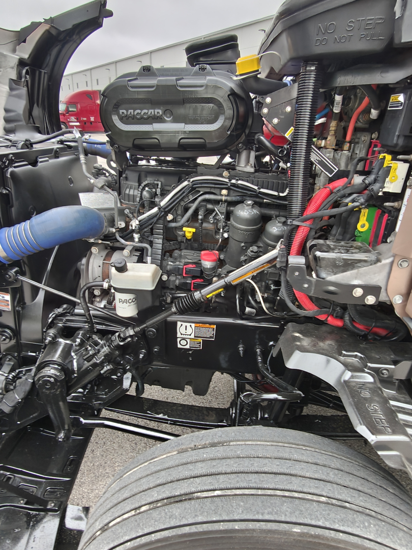 Kenworth T680 engine bay showing compact diesel engine with RDCAP labeled air intake, hoses, wiring, and exposed frame near front wheel.