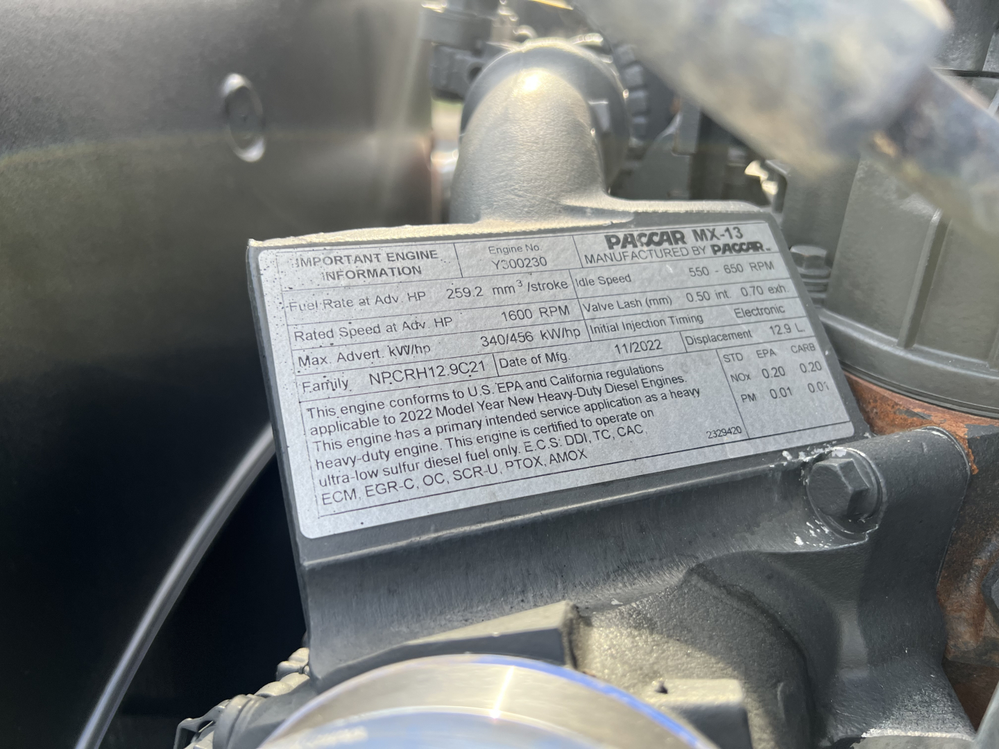 Label on engine cover showing technical specs and warnings for engine model PacCAR MX-13 in a Kenworth T680, with adjacent hoses and metal components.
