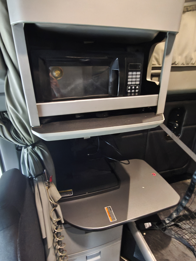 Cab interior of a Kenworth T680 showing a mounted microwave in a shelf and a folded desk/console with a control pad; no readable model text visible.