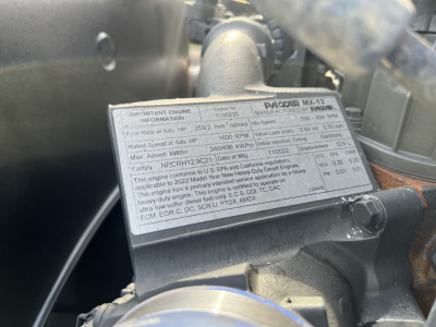 Label on engine cover showing technical specs and warnings for engine model PacCAR MX-13 in a Kenworth T680, with adjacent hoses and metal components.