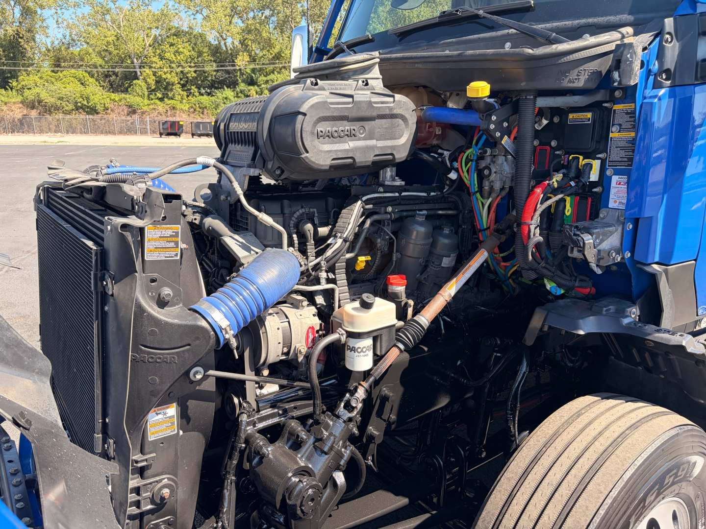 Kenworth T680 engine bay with V8 Cummins/Detroit engine visible and various hoses and wiring harnesses. Radiator core support on left, blue intake hose, yellow oil cap, and exposed turbo area.