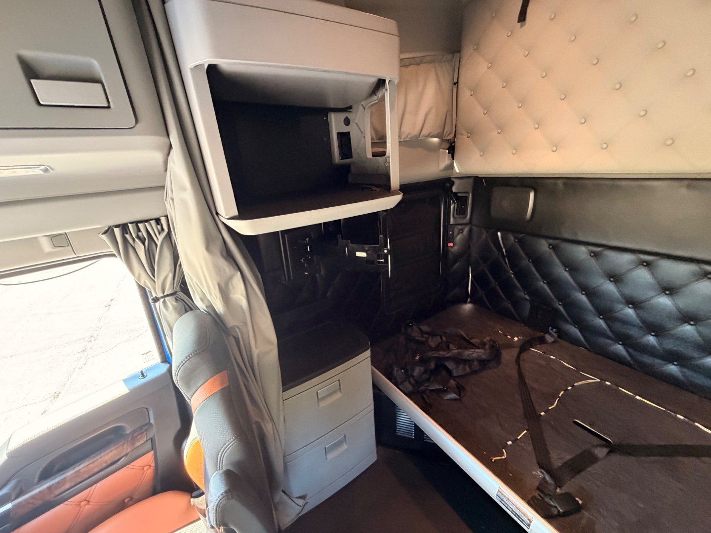 Cab interior of a Kenworth T680 sleeper area with beige quilted wall paneling, driver seat, and open storage cabinet above the dash.