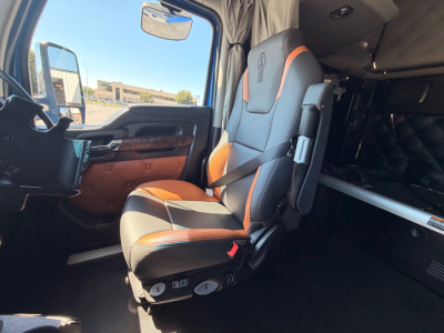 Truck cab seat in a 2023 Kenworth T680, driver seat with black and orange leather, dashboard area, side window, interior sleeper area visible.