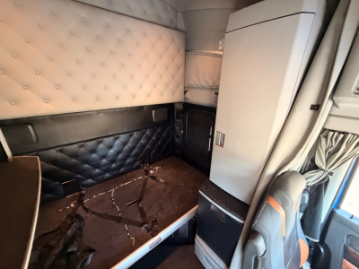 Kenworth T680 truck cabin interior showing sleeper area with tufted white upper wall and black lower wall, seat and storage cabinet visible, brown floor with straps.