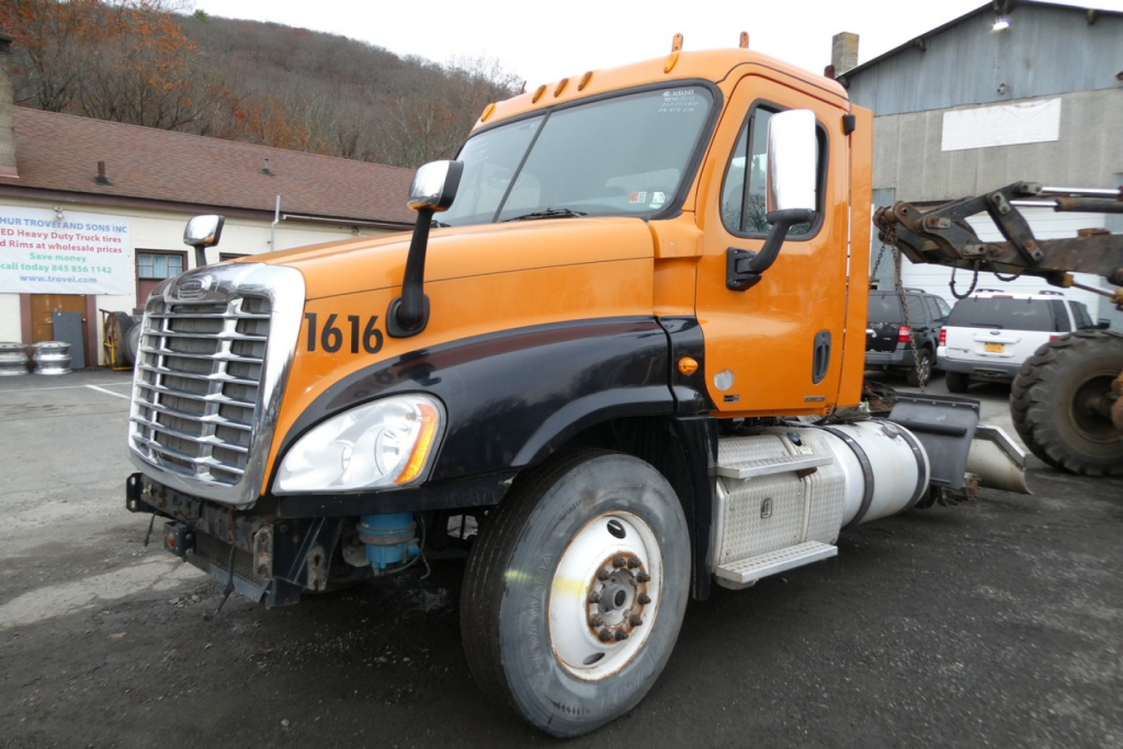 Truck side view showing Freightliner Cascadia 125 conventional daycab, orange cab, black lower fairing, visible front grille and wheel wells.
