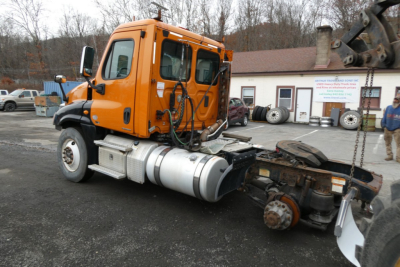 Orange Freightliner Cascadia 125 daycab truck tractor seen from side, chassis and fuel tanks visible, rear drive axle exposed, VIN plate and stock number in background.