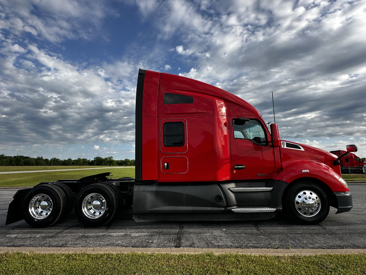 Kenworth T680 side view showing sleeper cab, red exterior, chrome wheels, three rear axles, and fuel/tank area.