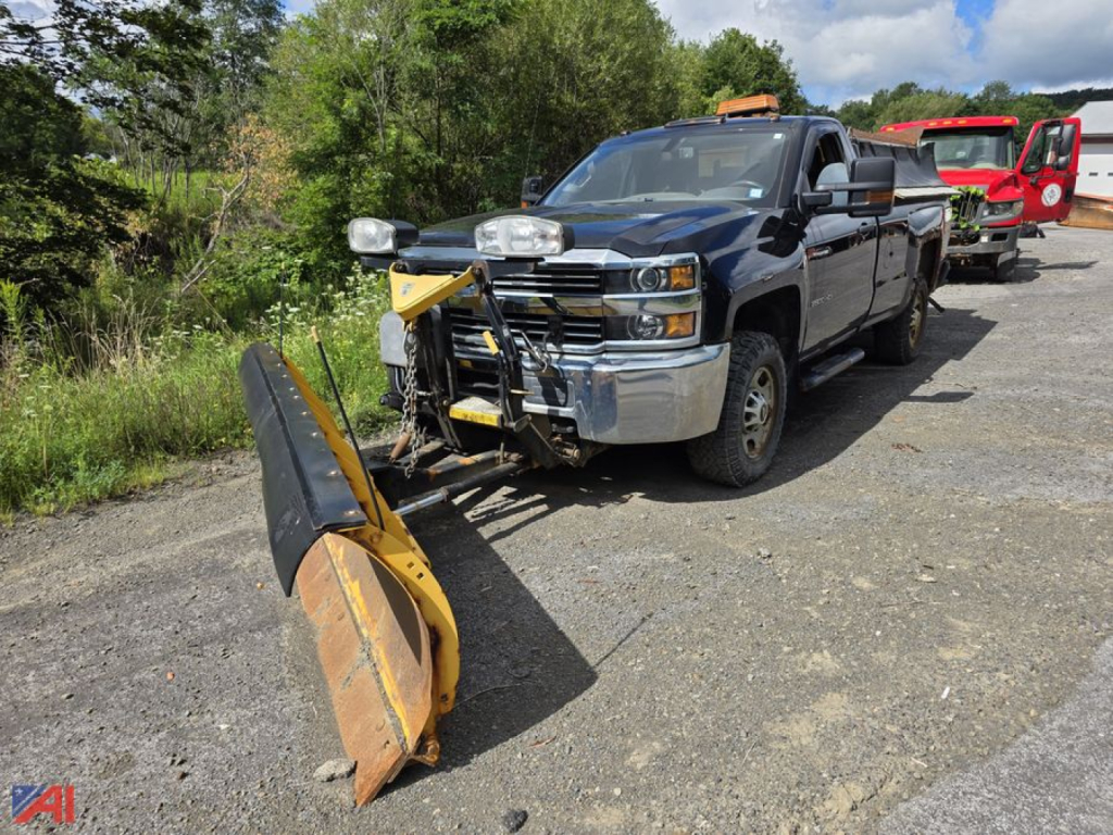 Chevrolet Silverado 2500HD pickup with snow plow and sander attachment parked on gravel; front chrome bumper, black crew cab, and yellow snow plow in foreground.