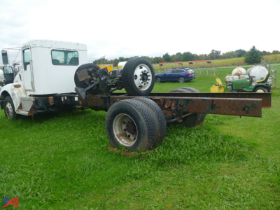 Kenworth T300 chassis cab forward view showing bare frame and rear dual wheels, front cab with a white exterior, overcast field in background.