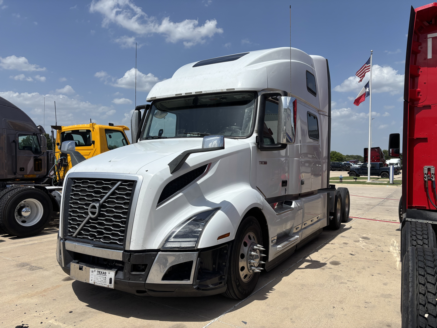 Volvo VNL64T860 highway truck in a lot, front-left view, with white cab and sleeper, chrome grille, side mirrors, and a small American flag in background.